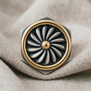 May include: A hexagonal ring featuring a central circular motif with radiating silver-toned fan-like blades, surrounded by a smooth gold-toned border. The center of the fan is accented with a small, polished gold-toned sphere, set against a dark background.