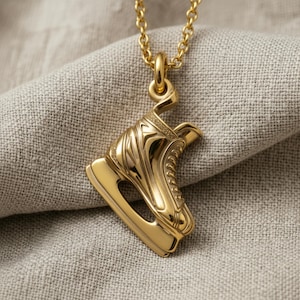 Op de afbeelding: Een goudkleurige metalen hanger in de vorm van een schaats, hangend aan een bijpassende goudkleurige ketting. De hanger is voorzien van gedetailleerde, gegoten texturen die de schoen, de veters en het ijzer van de schaats weergeven.