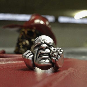 May include: A silver ring with a sculpted face, furrowed brow, and clenched fists. The word "HOOLIGAN" is engraved on the ring. The ring is set against a red surface, with a blurred, dark red object in the background.