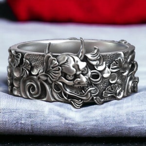 May include: Silver ring with a detailed design of a Japanese demon face surrounded by floral patterns.