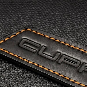 Op de afbeelding: Close-up van een zwart leren label met het in reli&euml;f aangebrachte woord "CUPRA" en oranje stiksels langs de randen. Het label staat tegen een achtergrond van zwart leer met textuur.