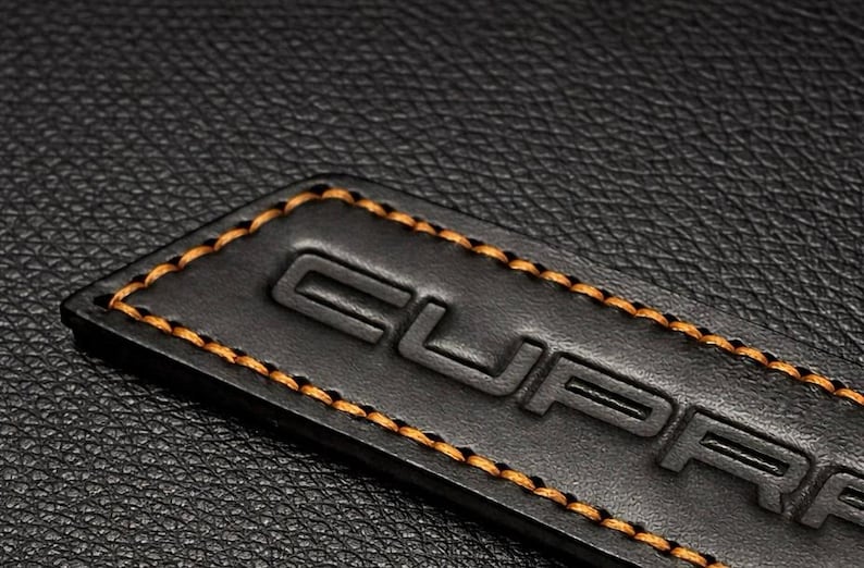 Op de afbeelding: Close-up van een zwart leren label met het in reli&euml;f aangebrachte woord "CUPRA" en oranje stiksels langs de randen. Het label staat tegen een achtergrond van zwart leer met textuur.