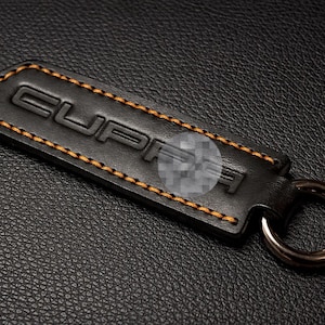 Op de afbeelding: Zwarte leren sleutelhanger met het woord "CUPRA" in reli&euml;f en oranje stiksels langs de randen. De sleutelhanger heeft een zilverkleurige metalen ring voor sleutels.