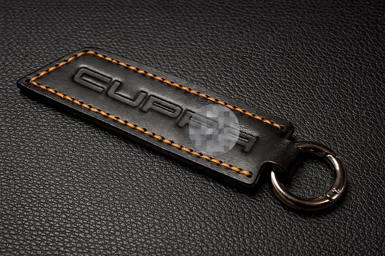 Op de afbeelding: Zwarte leren sleutelhanger met het woord "CUPRA" in reli&euml;f en oranje stiksels langs de randen. De sleutelhanger heeft een zilverkleurige metalen ring voor sleutels.