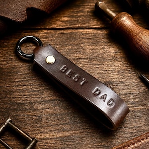 Può includere: Un portachiavi in pelle marrone scuro con il testo BEST DAD in rilievo. Il cinturino è fissato con un rivetto metallico color ottone e collegato a un anello brisé in metallo nero, creando un semplice design a occhiello per le chiavi.