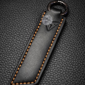 Op de afbeelding: Zwarte leren sleutelhanger met oranje stiksels en een zilveren sleutelring. De sleutelhanger heeft een geometrisch ontwerp en is gemaakt van getextureerd leer. De stiksels zijn zichtbaar langs de randen van de sleutelhanger.