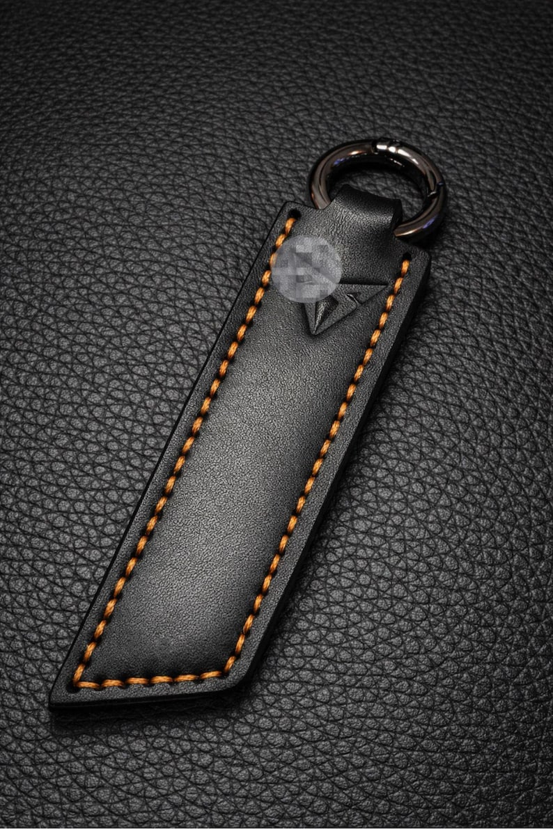 Op de afbeelding: Zwarte leren sleutelhanger met oranje stiksels en een zilveren sleutelring. De sleutelhanger heeft een geometrisch ontwerp en is gemaakt van getextureerd leer. De stiksels zijn zichtbaar langs de randen van de sleutelhanger.