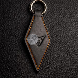 以下が含まれることがあります： 黒い革製のキーホルダー。ひし形をしており、金色のステッチが施されています。キーホルダーには金色のリングと中央に黒いロゴがあります。背景は、テクスチャ加工された黒い表面です。