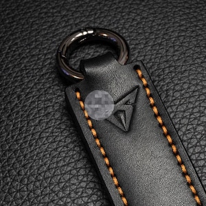 Op de afbeelding: Close-up van een zwart leren sleutelhanger met een zilverkleurige metalen ring. De sleutelhanger heeft oranje stiksels en een driehoekig reli&euml;f. De achtergrond is een zwarte, getextureerde ondergrond.