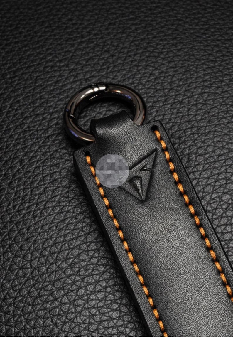 Op de afbeelding: Close-up van een zwart leren sleutelhanger met een zilverkleurige metalen ring. De sleutelhanger heeft oranje stiksels en een driehoekig reli&euml;f. De achtergrond is een zwarte, getextureerde ondergrond.