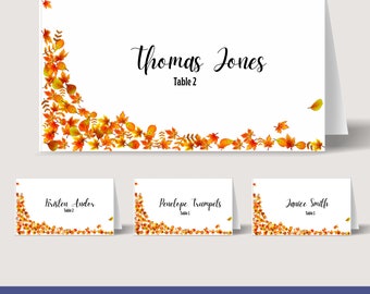 May include: Printable place cards with an autumn theme. The cards feature a white background with a border of orange and yellow leaves. The text "Thomas Jones Table 2" is printed on the top card. The other cards have the names "Kristen Andor Table 2", "Penelope Trumpets Table 1", and "Janice Smith Table 1".