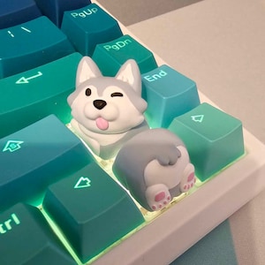 Könnte beinhalten: Ein weißer und grauer Husky-förmiger Keycap mit einer rosa Zunge, die herausragt, auf einer blaugrünen und weißen Tastatur.