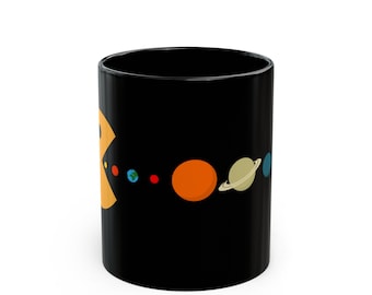 May include: A black ceramic mug featuring a graphic of a yellow, circular character with an open mouth positioned to the left of a horizontal line of colorful planets, including Earth, Mars, Jupiter, and Saturn, arranged across the surface.