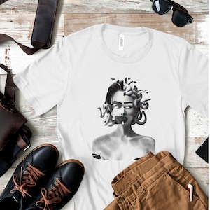 May include: A white short-sleeve t-shirt featuring a black and white graphic print of a split face. One side shows a human face, while the other side depicts Medusa with snakes for hair. A black rectangular bar covers the mouth area.