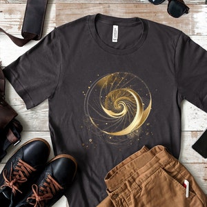 Könnte beinhalten: Ein dunkelgraues T-Shirt mit einem goldenen Spiralmuster auf der Vorderseite. Die Spirale ist von einem Kreis aus kleineren Punkten umgeben. Das Shirt liegt flach auf einer Holzoberfläche.