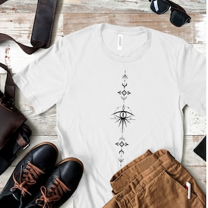 May include: White t-shirt with a black and white geometric design featuring an eye, moons, and stars. The design is centered on the shirt and extends vertically down the front.