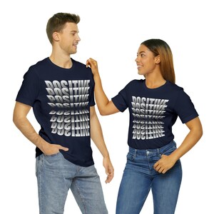 May include: Two people wearing navy blue t-shirts with a silver "POSITIVE" graphic printed on the front. The graphic is repeated multiple times, with each repetition slightly offset from the previous one, creating a layered effect.