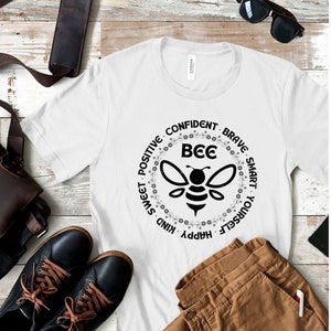 Peut inclure: T-shirt blanc avec un graphique d'abeille noir et blanc et le texte "Bee" au centre d'un cercle. Le cercle est entouré des mots "Happy, Kind, Sweet, Positive, Confident, Brave, Smart, Yourself".