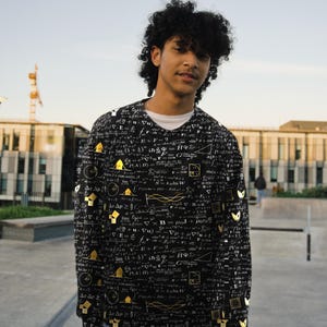 May include: A black long-sleeve shirt featuring a repeating white and gold print of various mathematical equations, geometric shapes, and scientific symbols, worn over a white crew neck undershirt with blue denim jeans.