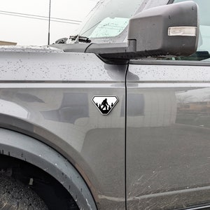 Puede incluir: Un coche gris con una pegatina en blanco y negro de la silueta de un Bigfoot en forma de diamante en el lateral.