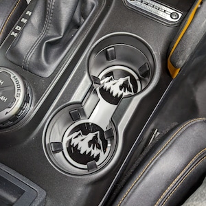 Op de afbeelding: Twee zwarte bekerhouders met een bergketenontwerp in een auto-interieur. De bekerhouders zijn gemaakt van metaal en hebben een zwarte afwerking. Het auto-interieur is van zwart leer met gele stiksels.