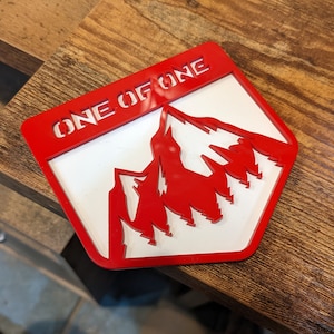 Puede incluir: Un letrero de plástico rojo y blanco con el texto "ONE OF ONE" y una silueta de montaña roja.