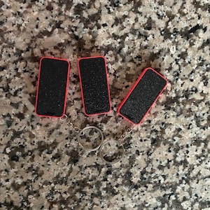 Puede incluir: Tres pequeños llaveros rectangulares con una superficie negra texturizada y un borde rojo. Los llaveros están conectados por una cadena plateada y sujetos a un llavero plateado. Los llaveros se muestran sobre una superficie gris moteada.