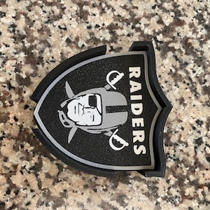 Puede incluir: Posavasos con el logotipo de los Raiders en negro y gris. El posavasos presenta el logotipo del equipo con la palabra "RAIDERS" en blanco. El posavasos tiene forma de escudo y un borde negro.