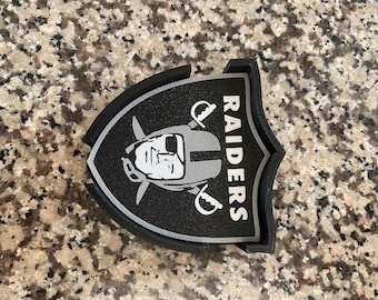 Puede incluir: Posavasos con el logotipo de los Raiders en negro y gris. El posavasos presenta el logotipo del equipo con la palabra "RAIDERS" en blanco. El posavasos tiene forma de escudo y un borde negro.