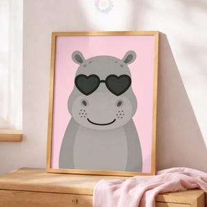 May include: A framed print featuring a cartoon hippo wearing heart-shaped sunglasses against a pink background. The hippo is gray with a smiling face. The frame is a light brown color. The print is displayed on a wooden dresser.
