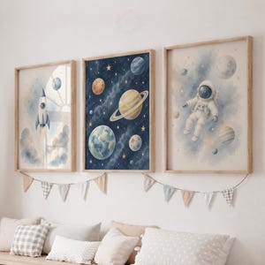 May include: Three framed wall art prints featuring space themes. The left print shows a rocket ship, the centre print displays planets and stars on a dark blue background, and the right print depicts an astronaut floating in space with planets.