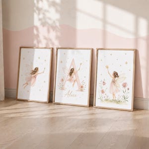 Può includere: Tre stampe da parete incorniciate con illustrazioni ad acquerello di fate con ali e bacchette magiche. La stampa centrale mostra una grande lettera A con il nome Alice scritto sotto. I disegni includono fiori rosa tenue e stelle dorate.