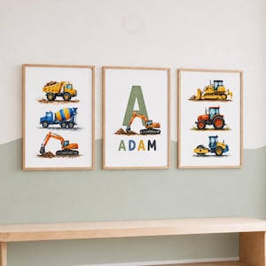 May include: Three framed wall art prints featuring watercolour illustrations of construction vehicles. The left print shows a yellow dump truck, blue cement mixer, and orange excavator. The centre print displays a large letter A with an excavator and the name ADAM. The right print shows a yellow bulldozer, orange tractor, and blue road roller.