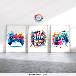 May include: Three framed art prints with a gaming theme. Each print features a colourful design: a video game controller, a controller with paint splatter, and the text "EAT SLEEP GAME REPEAT" with a paint splatter background. The prints are displayed on a white wall.