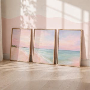 May include: A set of three rectangular wall art prints featuring a continuous coastal landscape. Each print displays a sandy beach meeting turquoise ocean waters under a soft pink and peach sunset sky, framed in light-coloured wooden borders.