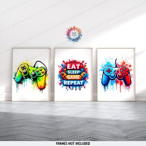 May include: Three framed prints with a gaming theme. The left print features a green and yellow video game controller with colorful paint splatters. The middle print has the text "Eat Sleep Game Repeat" in colorful letters with paint splatters. The right print features a red and blue video game controller with colorful paint splatters.