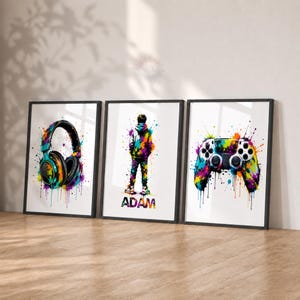May include: Three framed wall art prints featuring vibrant, multicoloured watercolour splatter designs. The left print shows headphones, the centre print depicts a person standing with the name ADAM below, and the right print displays a game controller.