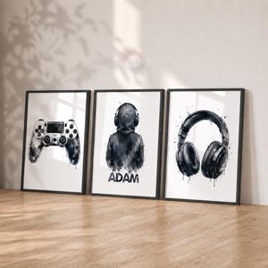 May include: Three framed wall art prints featuring black and white watercolour-style illustrations. The left print shows a game controller, the centre print depicts a hooded figure wearing headphones with the name ADAM, and the right print shows headphones.