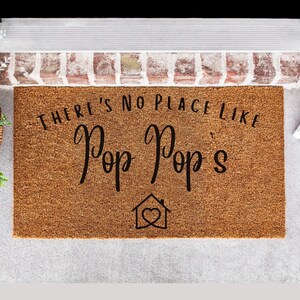 May include: A brown coir doormat with the text "There's no place like Pop Pop's" in black cursive lettering. A small house icon with a heart inside is below the text.