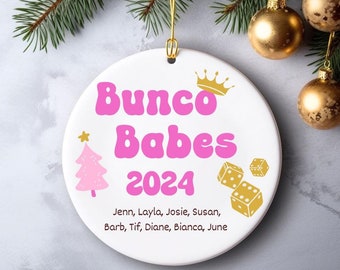 May include: A white ceramic ornament with the text "Bunco Babes 2024" in pink, along with a pink Christmas tree, a gold crown, and gold dice. The names of the group members are listed below the year. Gold hanging string.
