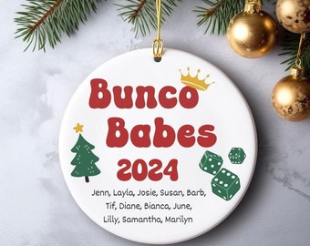 May include: White ceramic ornament with the text "Bunco Babes 2024" in red, a gold crown, a green Christmas tree, and green dice. The names of the Bunco Babes are listed below the year. Gold hanging string and gold Christmas ornaments are in the background.