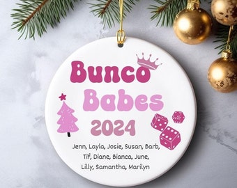 May include: White ceramic ornament with the text "Bunco Babes 2024" in pink, a pink Christmas tree, dice, and a crown. The names of the Bunco Babes are listed below the text. Gold hanging string and gold ornaments are in the background.