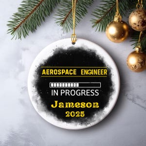 May include: A white ceramic Christmas ornament with a black and gray design. The text reads "AEROSPACE ENGINEER IN PROGRESS Jameson 2025" in yellow. Gold Christmas ornaments and pine branches are in the background.