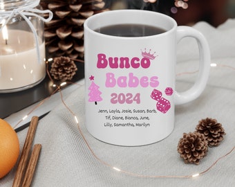 May include: A white ceramic mug featuring pink text that reads Bunco Babes 2024, accompanied by a pink crown, a pink Christmas tree, and pink dice. Below the text, a list of names is printed: Jenn, Layla, Josie, Susan, Barb, Tif, Diane, Bianca, June, Lilly, Samantha, Marilyn.