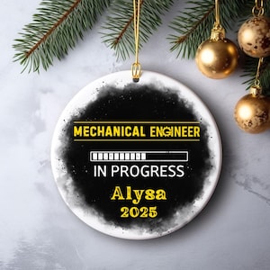 May include: A round, white ceramic ornament with the text "MECHANICAL ENGINEER IN PROGRESS Alysa 2025" in yellow. The design features a black and gray smoky background. Gold hanging hardware and Christmas ornaments are visible.