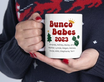 May include: White ceramic mug featuring the text "Bunco Babes 2023" in red, with a gold crown and green dice graphics. The mug also lists names. A green Christmas tree graphic and a gold star are also present.