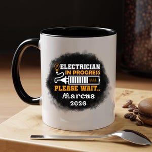May include: White ceramic mug with a black interior and handle. The mug features the text "ELECTRICIAN IN PROGRESS PLEASE WAIT.. Marcus 2026" in a black and orange design. A spoon rests on the wooden surface.