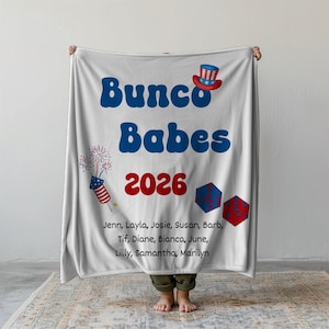 May include: White fleece blanket with the text "Bunco Babes 2026" in blue and red. The blanket also features a red, white, and blue top hat, fireworks, and dice. The names Jenn, Layla, Josie, Susan, Barb, Tif, Diane, Bianca, June, Lilly, Samantha, and Marilyn are listed below the main text.