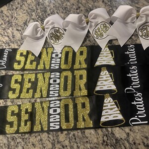 May include: Black satin sashes with gold glitter lettering that reads "SENIOR 2025" and personalized names. White bows with gold monogrammed initials are attached. Cheerleader megaphone graphics are also present.