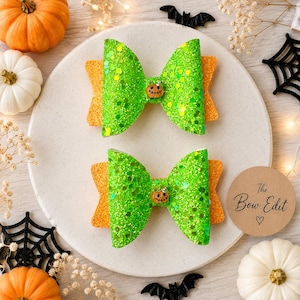 May include: Two hair bows featuring a bright green glitter finish layered over a solid orange glitter base. Each bow is centred with a small, round, orange pumpkin charm with a black jack-o'-lantern face and a tiny green stem detail.